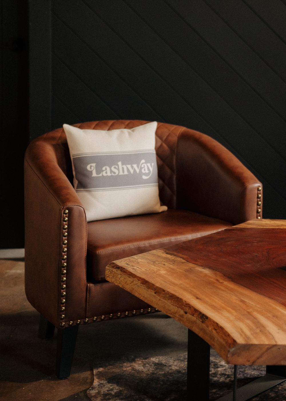 Brown leather barrel chair with Lashway branded pillow beside a live-edge wood coffee table against dark shiplap wall.