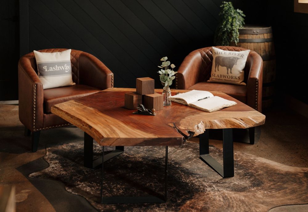 Live edge walnut coffee table with black metal legs on a cowhide rug, flanked by brown leather armchairs in a modern rustic living room.