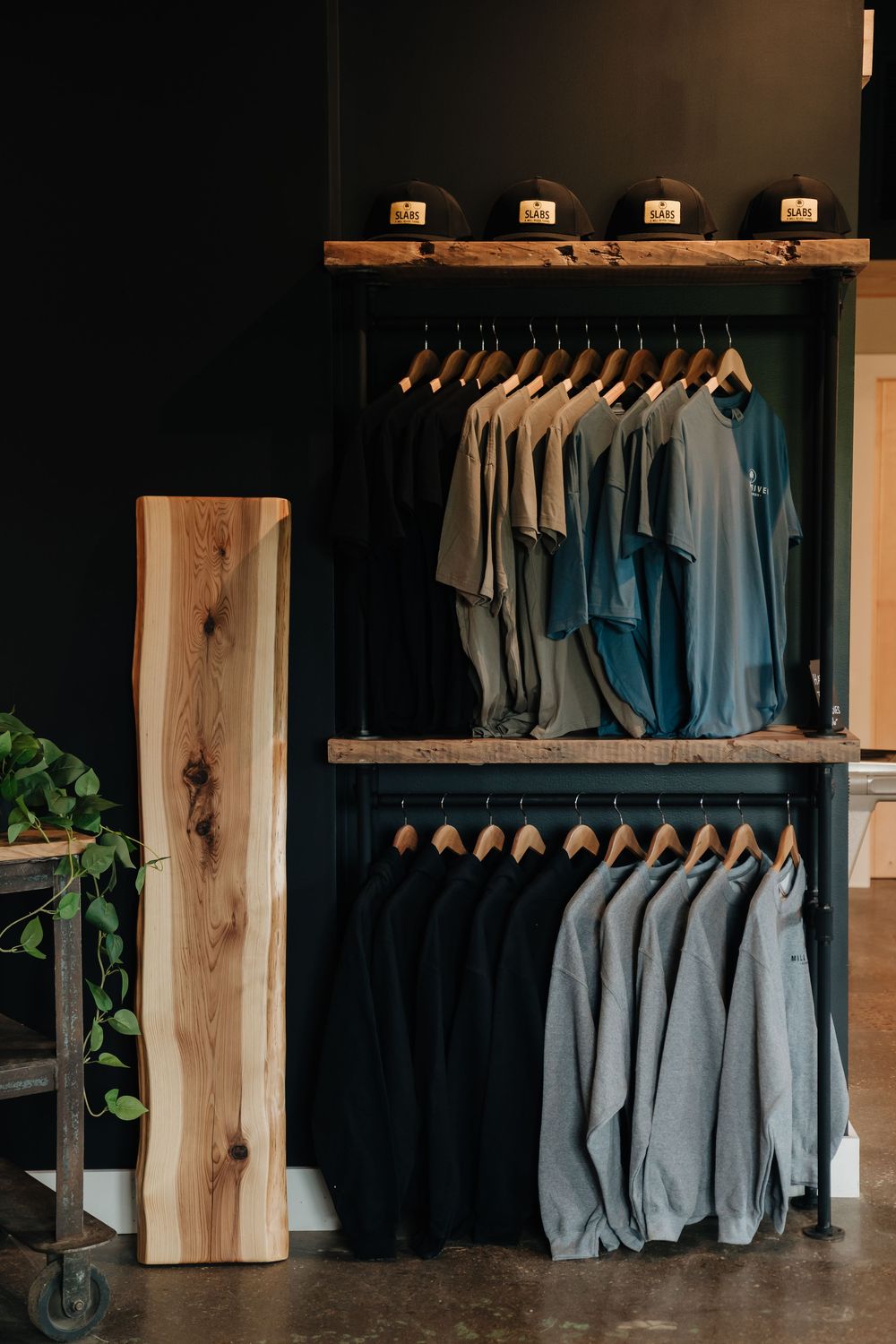 Rustic clothing store display with t-shirts on wooden shelves, hats on top, and hoodies on lower rack against dark walls.