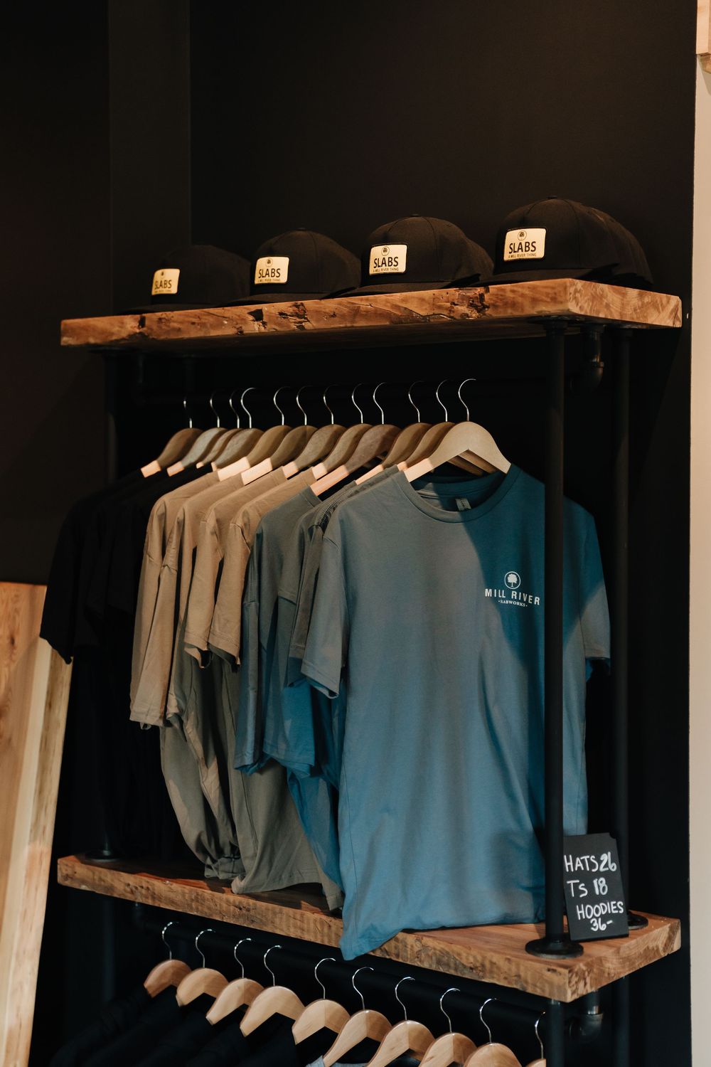 Rustic wooden clothing rack displaying gray and teal t-shirts on hangers, with black snapback hats on upper shelf.