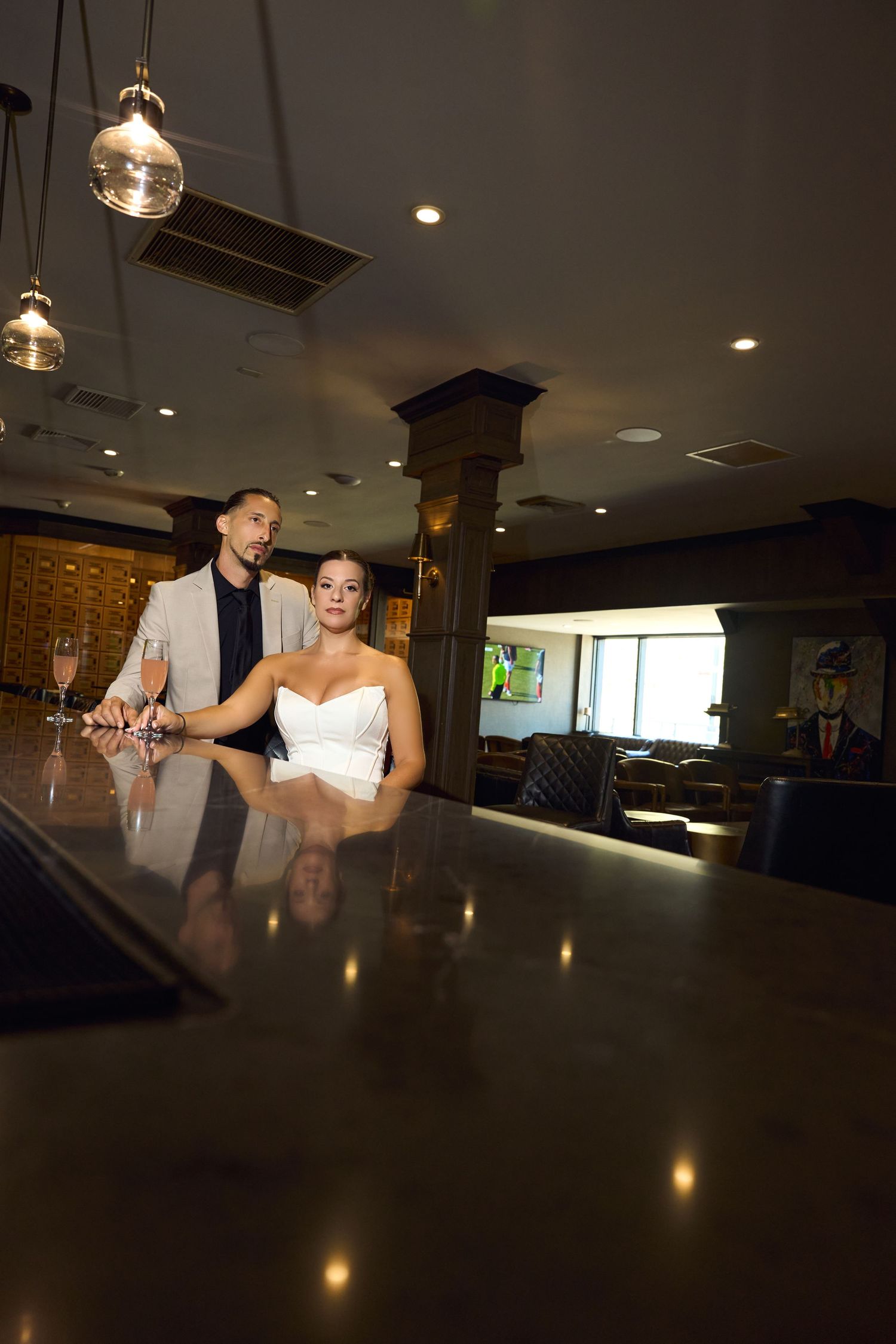 Bride in white dress and groom in suit pose at a dark wood bar with champagne glasses in an elegant venue.