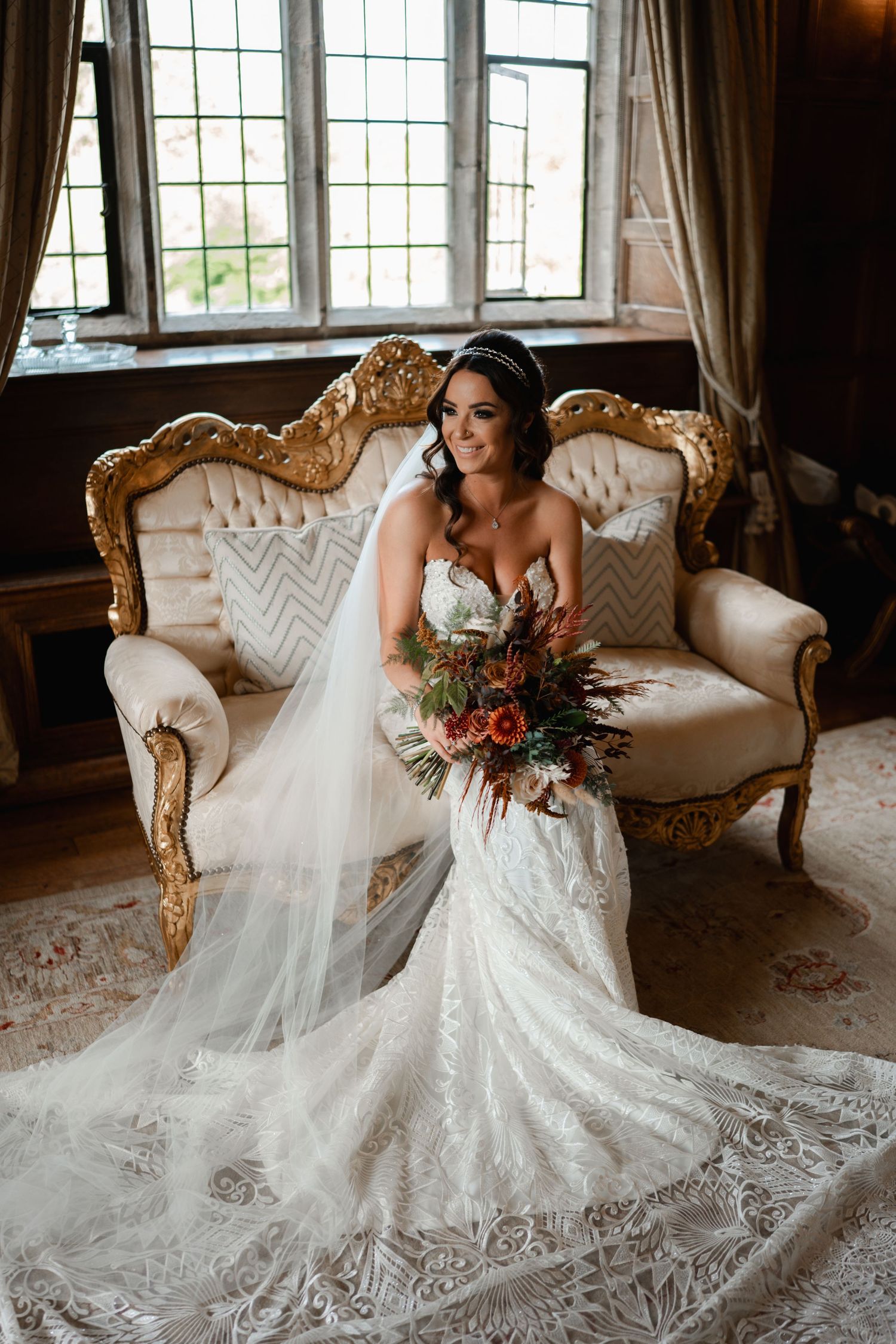 Bride in lace mermaid gown with veil sits on ornate gold sofa holding autumn bouquet by window.