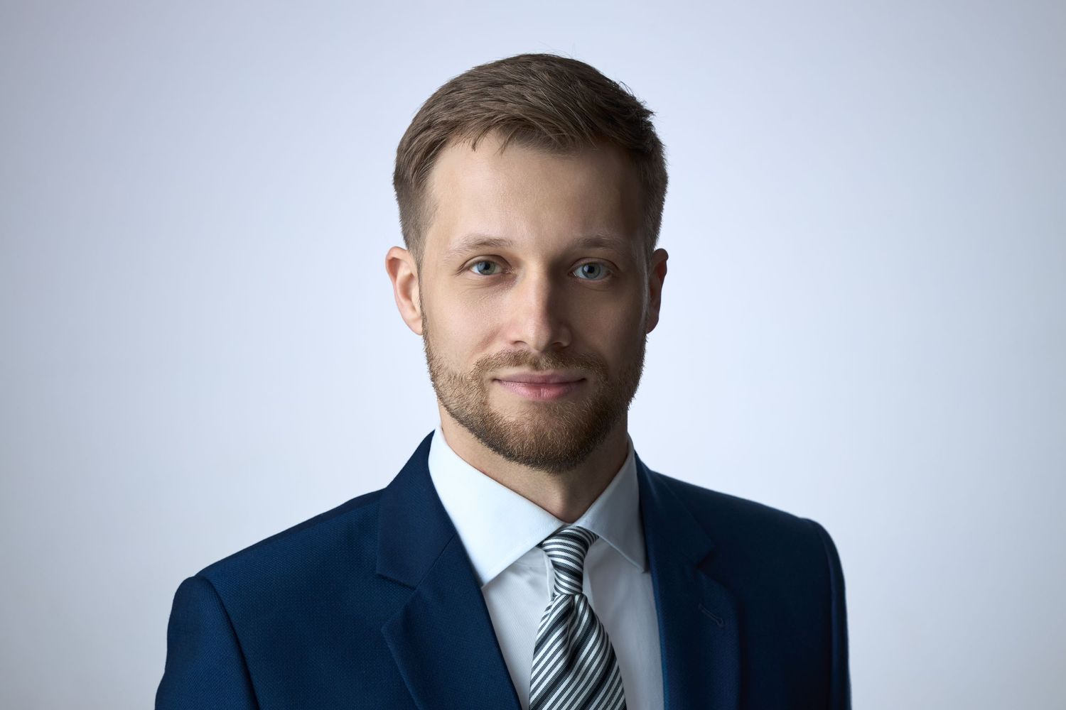 Professional young businessman in navy suit and striped tie smiling confidently against light gray background.