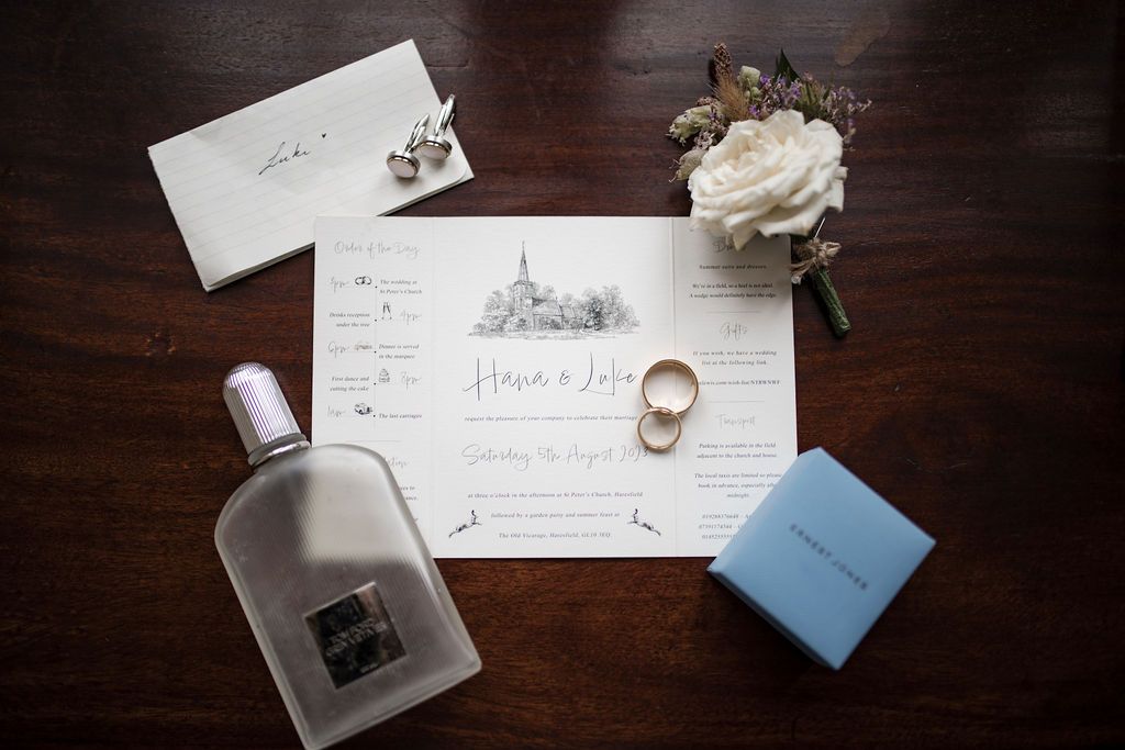 Elegant wedding flat lay featuring invitation suite, gold rings, white floral boutonniere, perfume bottle, and blue gift box on dark wood.