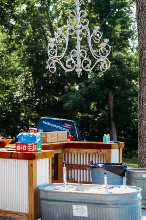 Outdoor rustic bar with corrugated metal, wooden countertop, beverages, galvanized tubs, and elegant white chandelier hanging above.