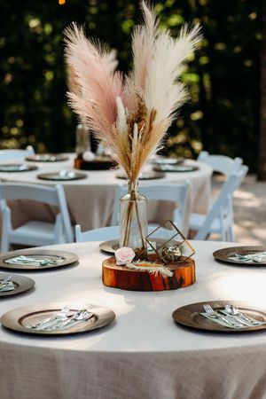 Boho wedding table with pampas grass centerpiece on wood slice, dark charger plates, and white linens outdoors.