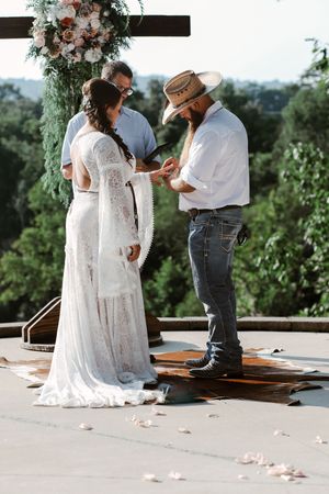 Bride and groom exchange rings outdoors at rustic wedding ceremony, groom wearing cowboy hat and boots.