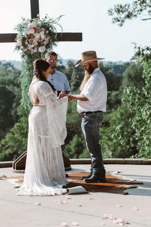 Boho bride and bearded groom exchange vows outdoors under floral cross arch with lush green trees backdrop.