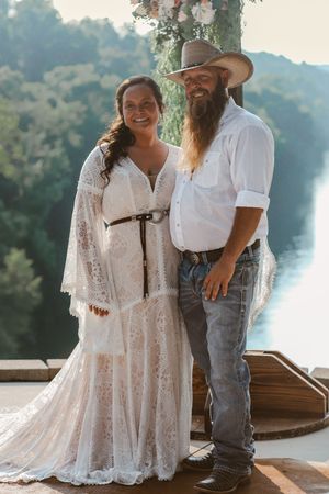Bride in lace boho gown and groom in cowboy hat and jeans pose together at outdoor riverside wedding ceremony.