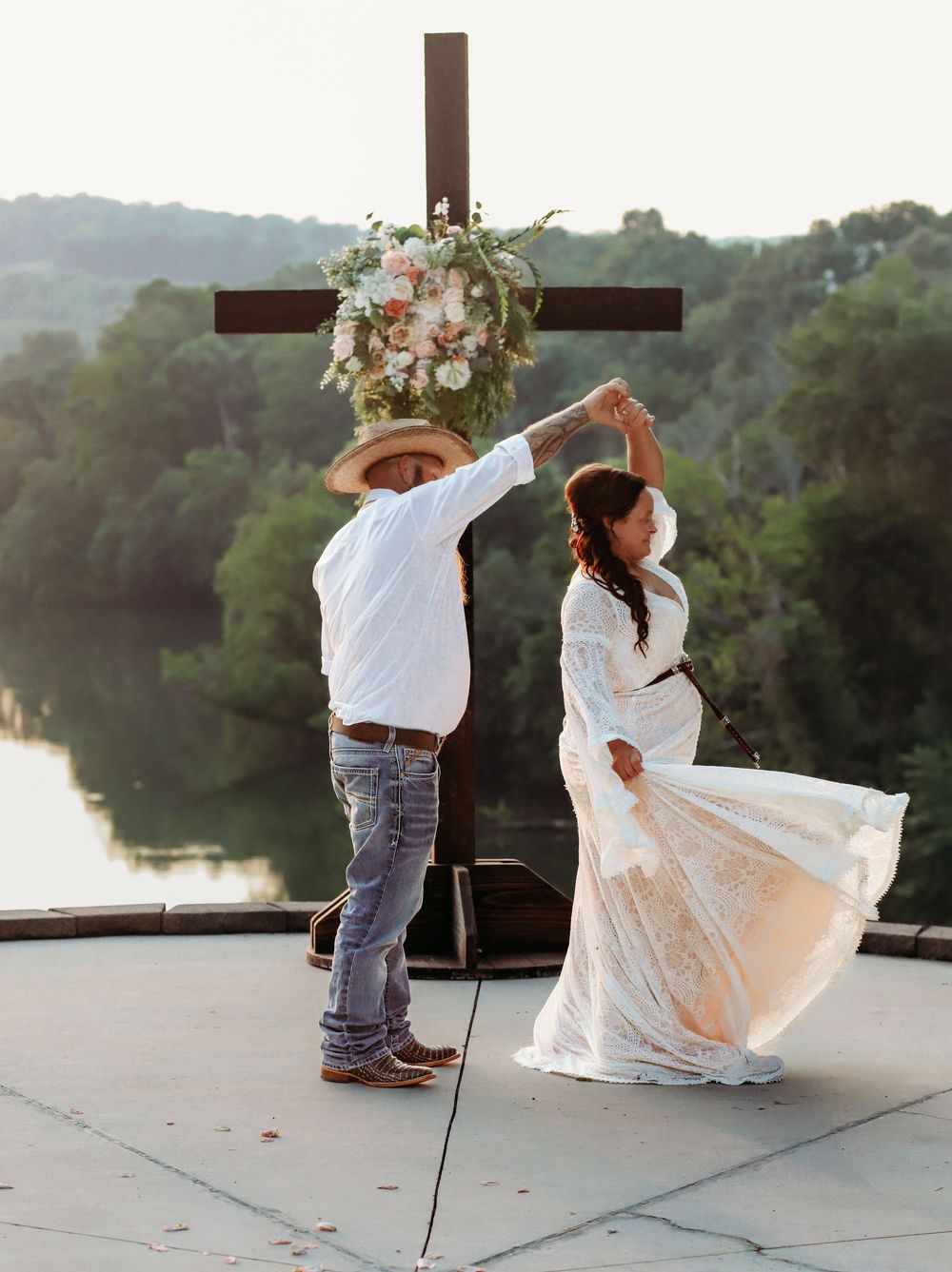 Couple dancing at outdoor wedding ceremony with cross and floral arrangement, scenic river and hills in background.