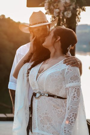 Couple in bohemian wedding attire sharing a romantic moment at golden hour by a lake, bride in lace dress.
