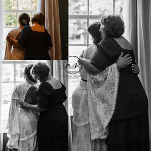 Black and white wedding photo of bride and older woman embracing warmly by a bright window in elegant lace gown.