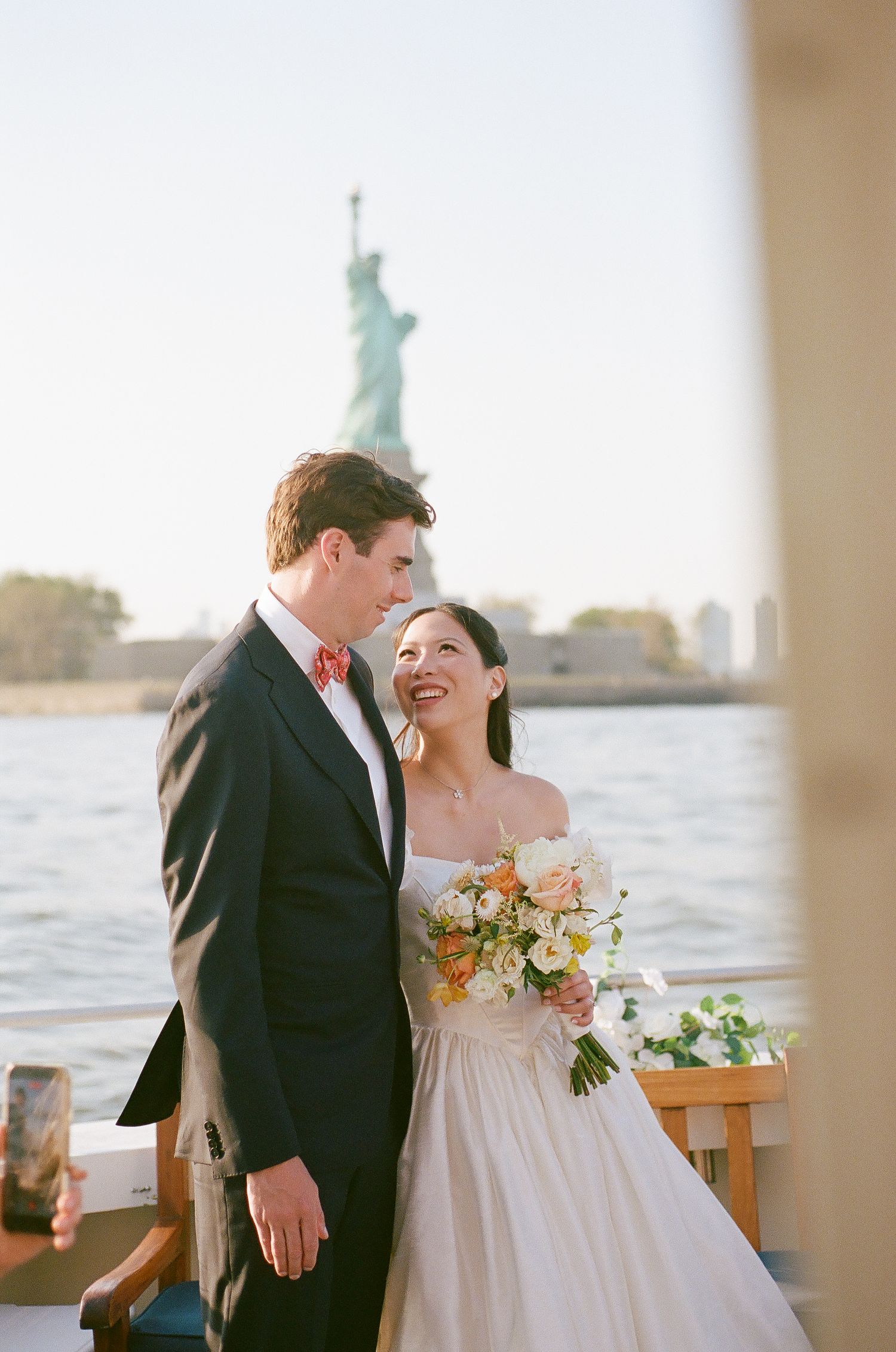 Bride and groom pose on a boat with the Statue of Liberty in the background on their wedding day.