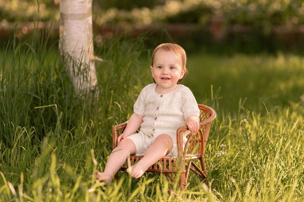 A smiling baby in a beige romper sits in a small wicker chair on lush green grass during childhood photoshoot in Lincolnshire.
