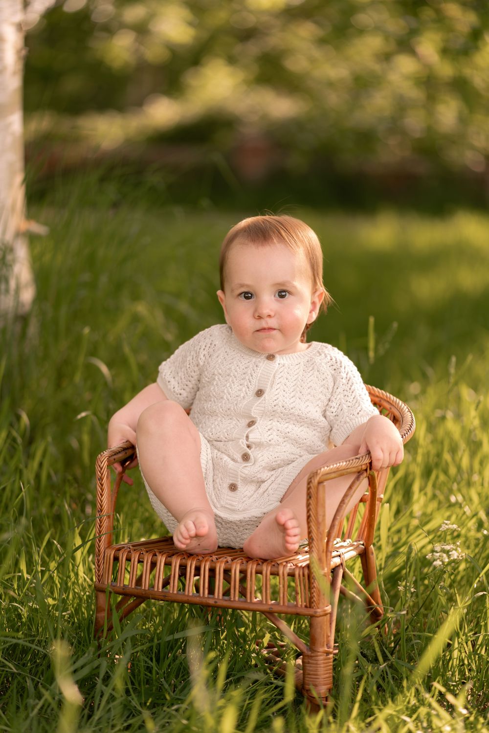 Adorable baby in cream knit romper sitting in small wicker chair on green grass outdoors during childhood photoshoot in Lincolnshire.