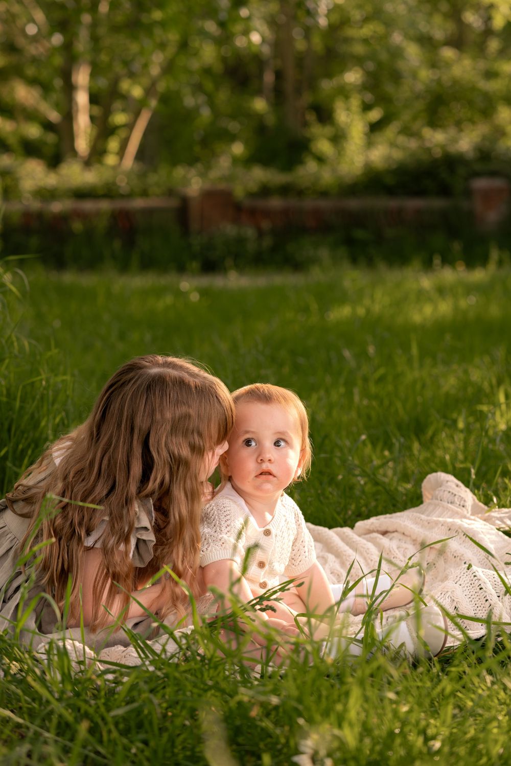 Older sister whispering to baby sibling lying on blanket in lush green grass during golden hour during childhood photoshoot in Lincolnshire.