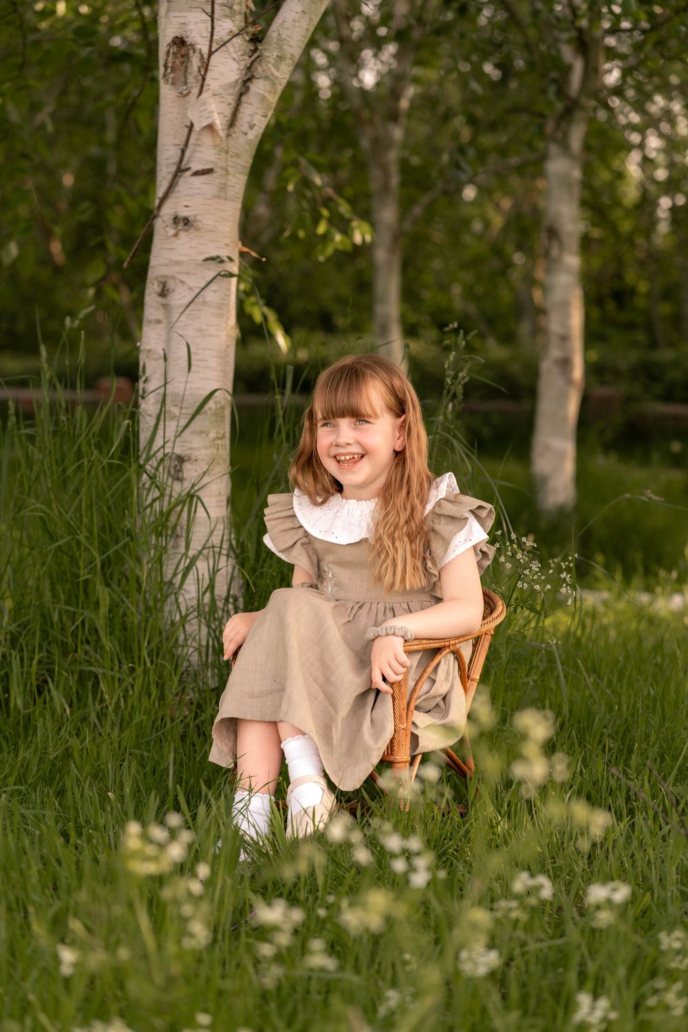 A young girl in a linen dress sits on a wicker chair among wildflowers beside birch trees during childhood photoshoot in Lincolnshire.