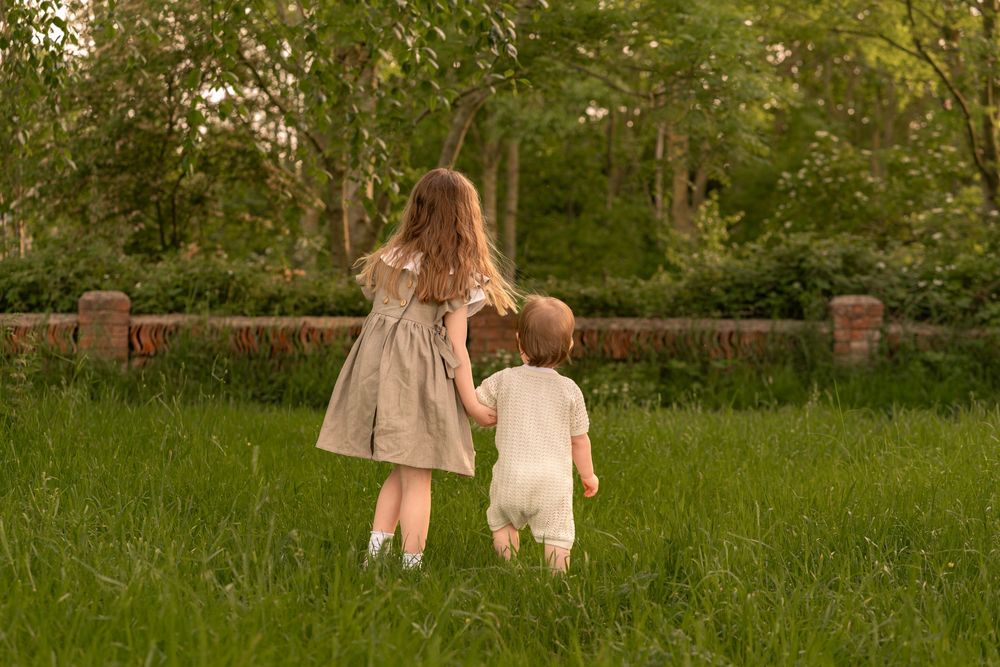 Two young children holding hands walking through lush green grass near trees on a sunny day during childhood photoshoot in Lincolnshire.