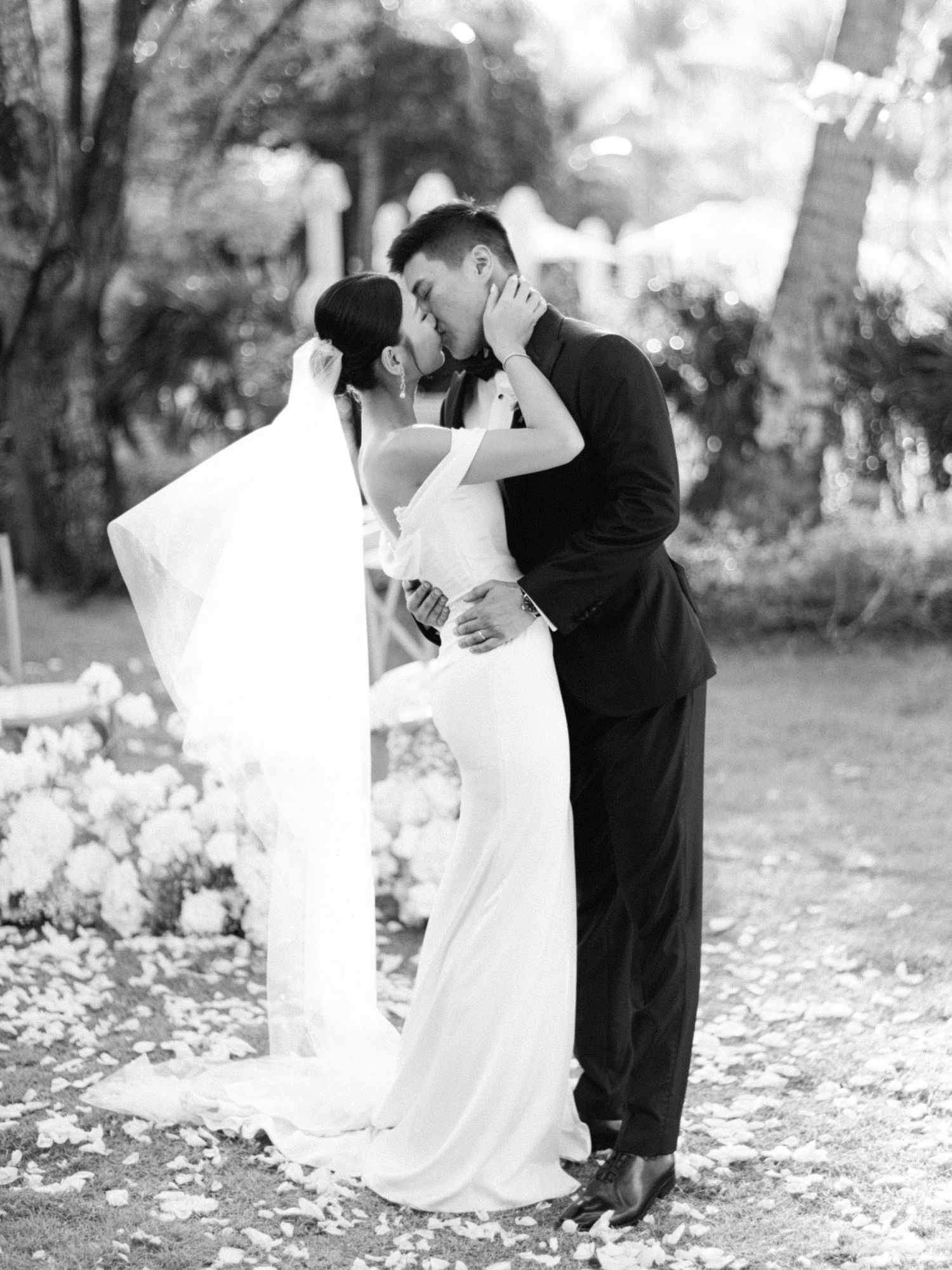 Black and white photo of a bride and groom sharing a first kiss at an outdoor wedding ceremony.