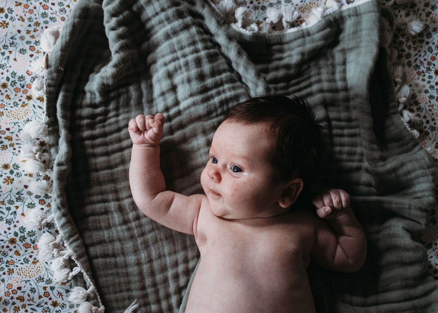Adorable baby lying on a soft gray muslin blanket with arms raised, looking up with curious eyes.
