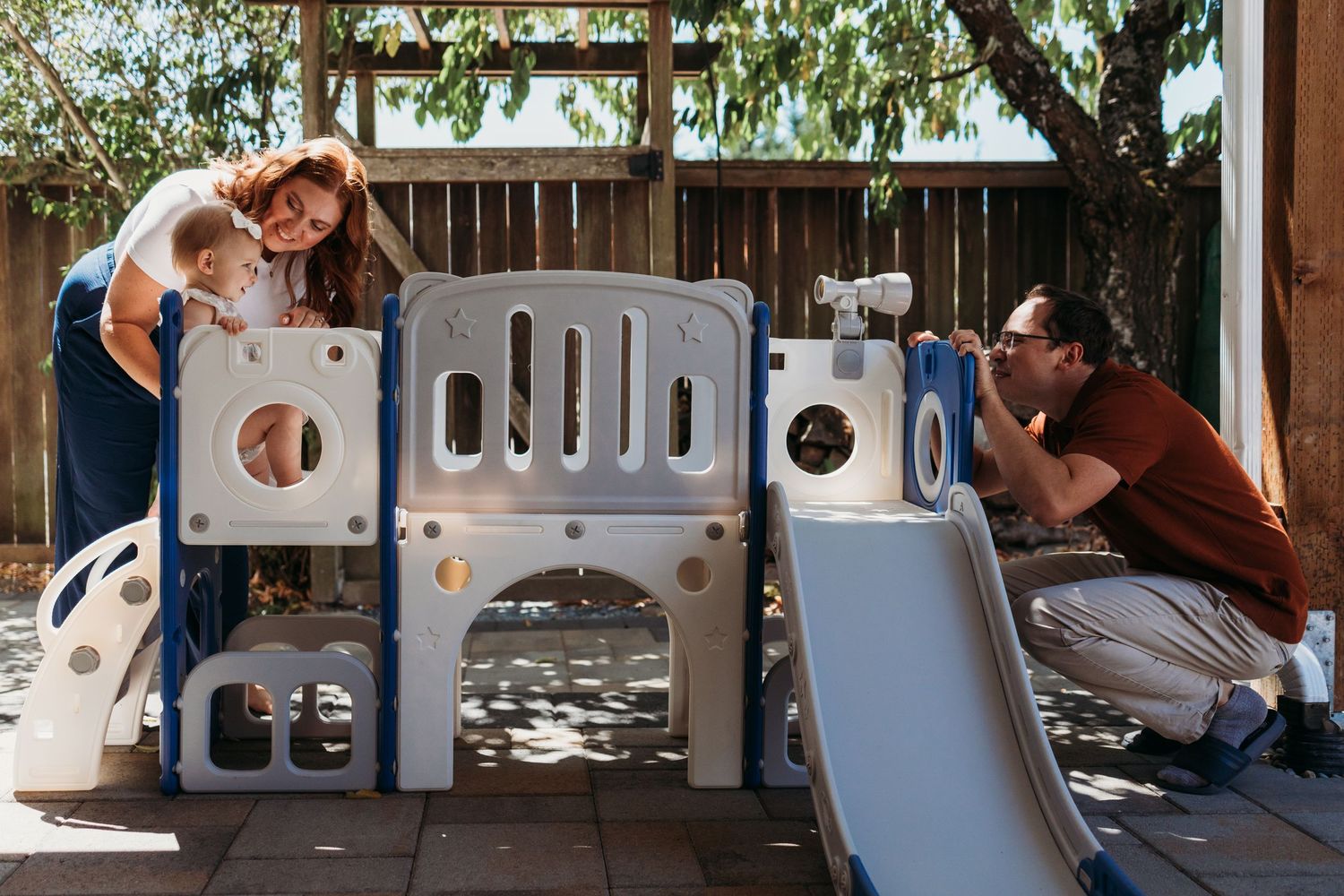 Family with toddler playing on outdoor plastic playground set in backyard patio area.