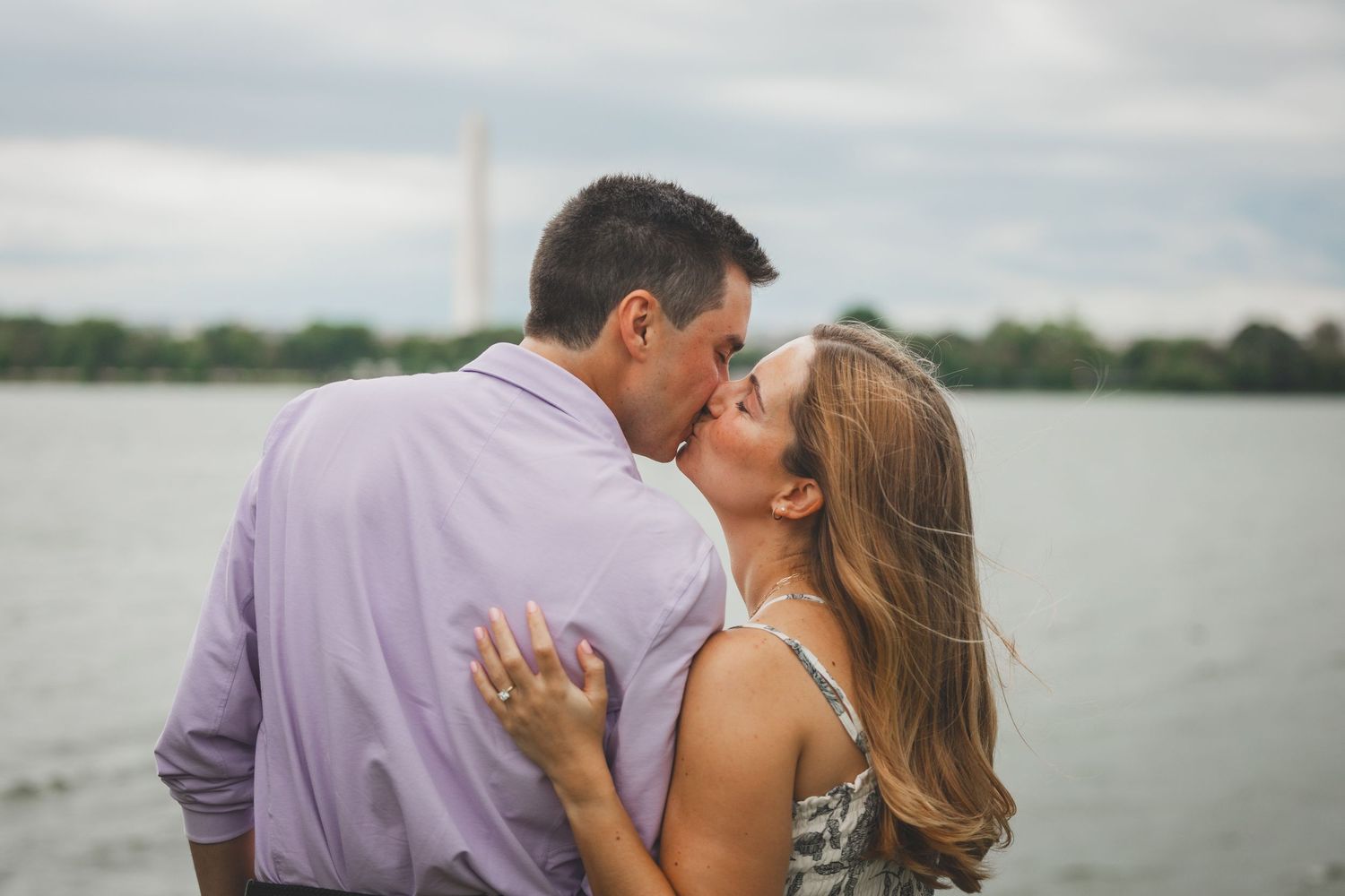 Couple sharing a romantic kiss near a waterfront with the Washington Monument visible in the background.