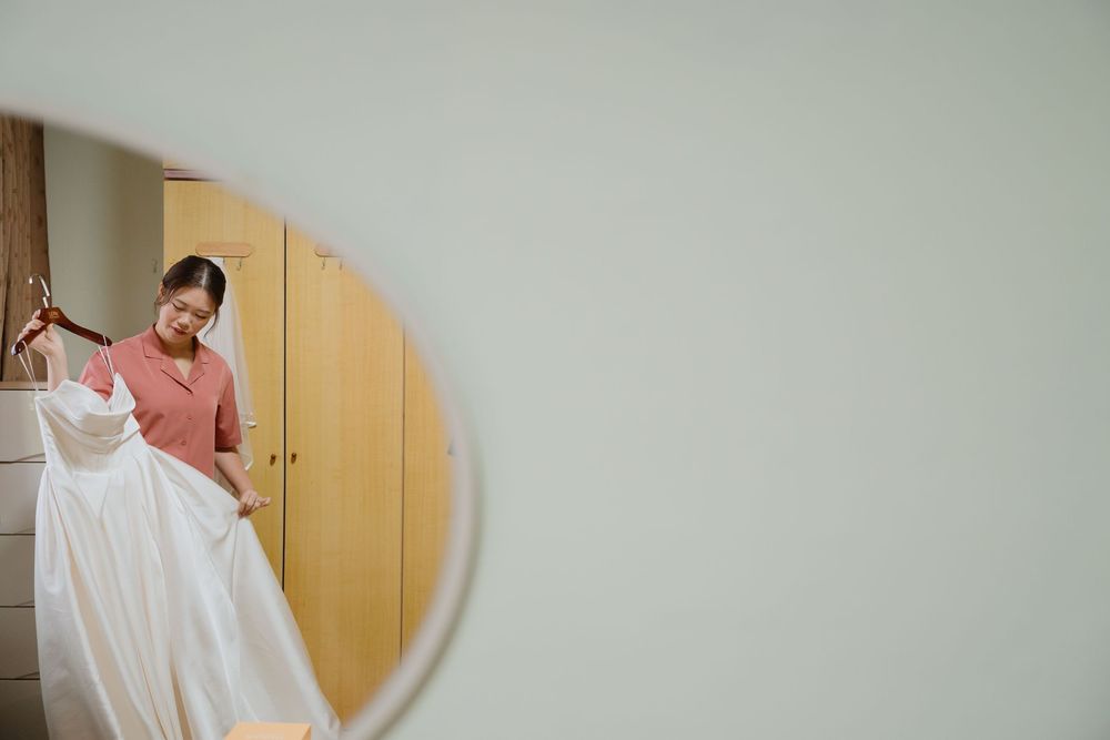 Woman in pink top holding a white wedding dress reflected in a round mirror in a bright room.