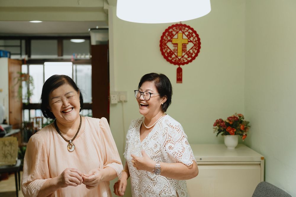 Two elderly Asian women laughing together indoors with a decorative Chinese wall ornament in the background.