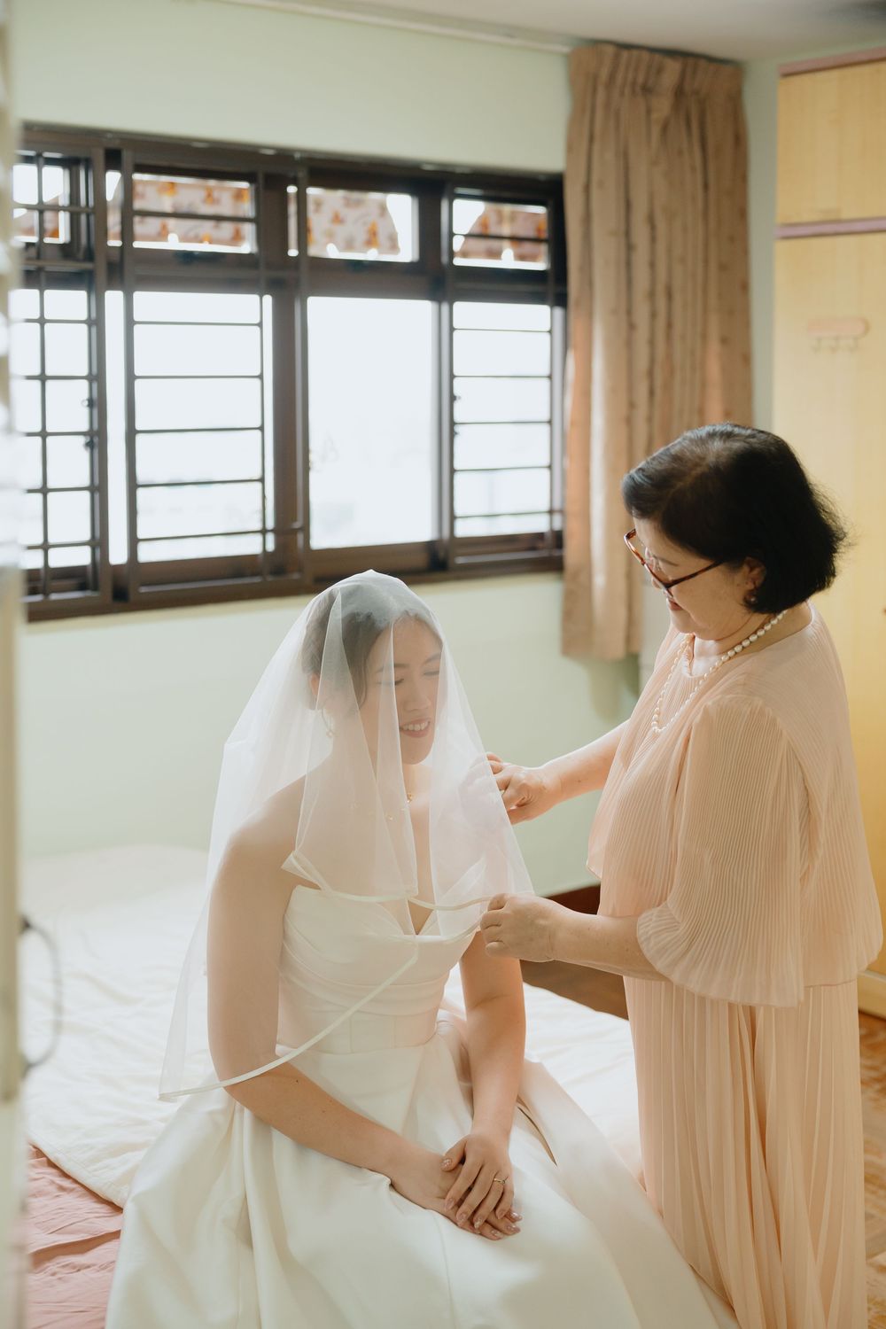 A mother helps her daughter put on a white bridal veil before the wedding ceremony in a softly lit room.
