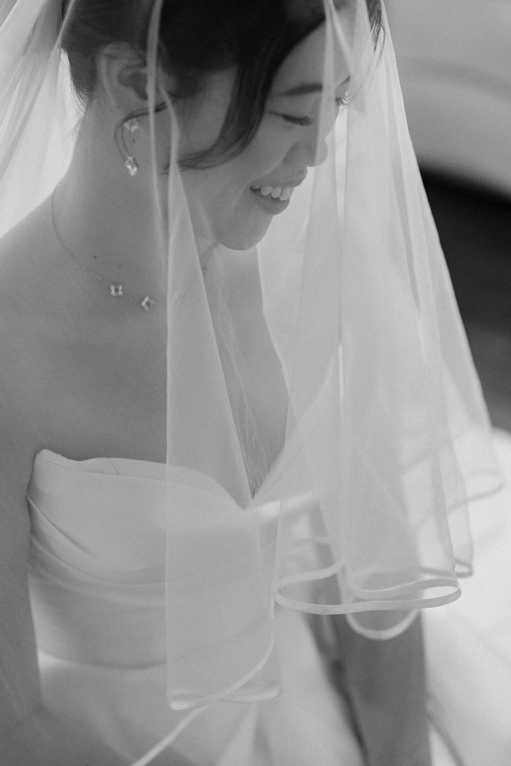 Black and white portrait of a smiling Asian bride in a strapless gown and veil, wearing delicate jewelry.