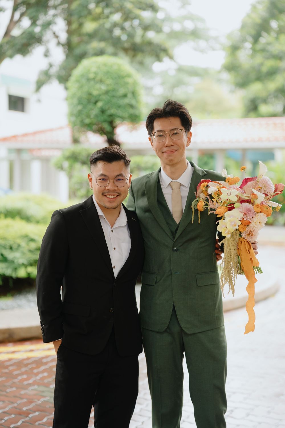 Two men in suits pose outdoors, one in black holding a colorful bouquet, the other in green, at a formal event.