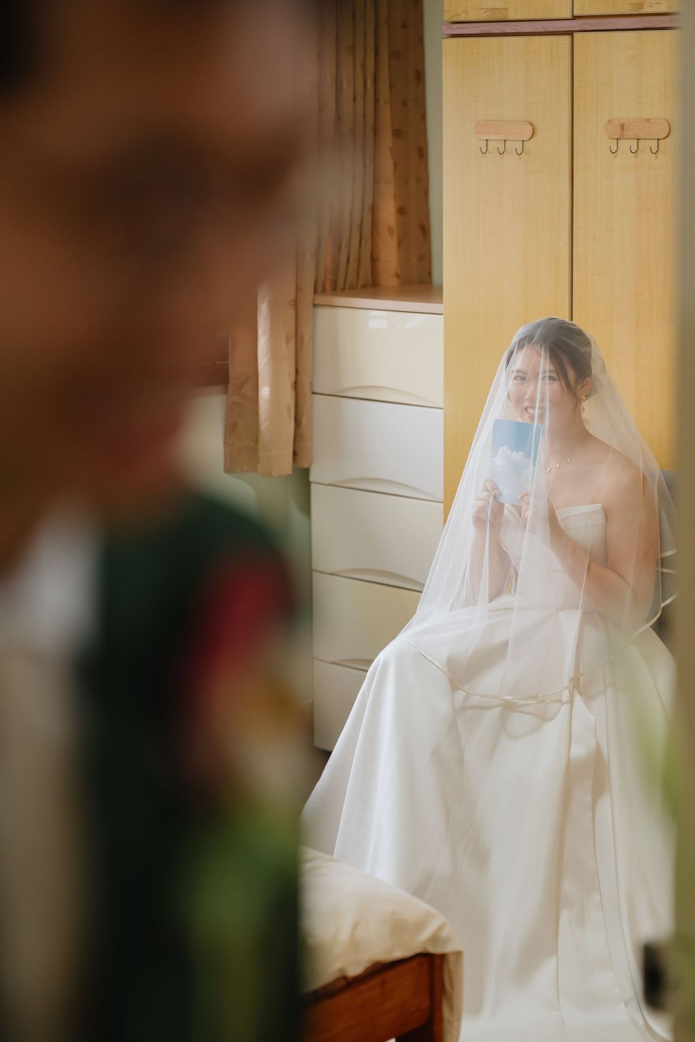 Bride in white wedding gown and veil sits in a room, wearing a blue face mask, reflected in a mirror.