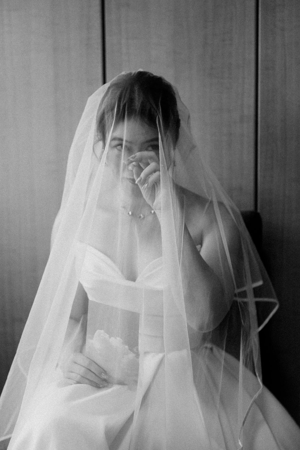 Black and white photo of a bride in a strapless gown, veil draped over her face, seated against a wooden backdrop.