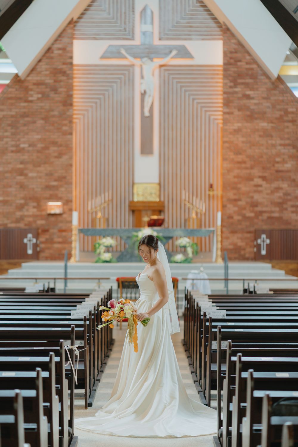 Bride in white gown holding colorful bouquet stands in church aisle with cross altar backdrop.