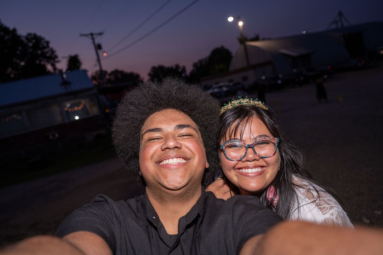 Two smiling friends take a selfie together outdoors at dusk, with lights and buildings visible in the background.