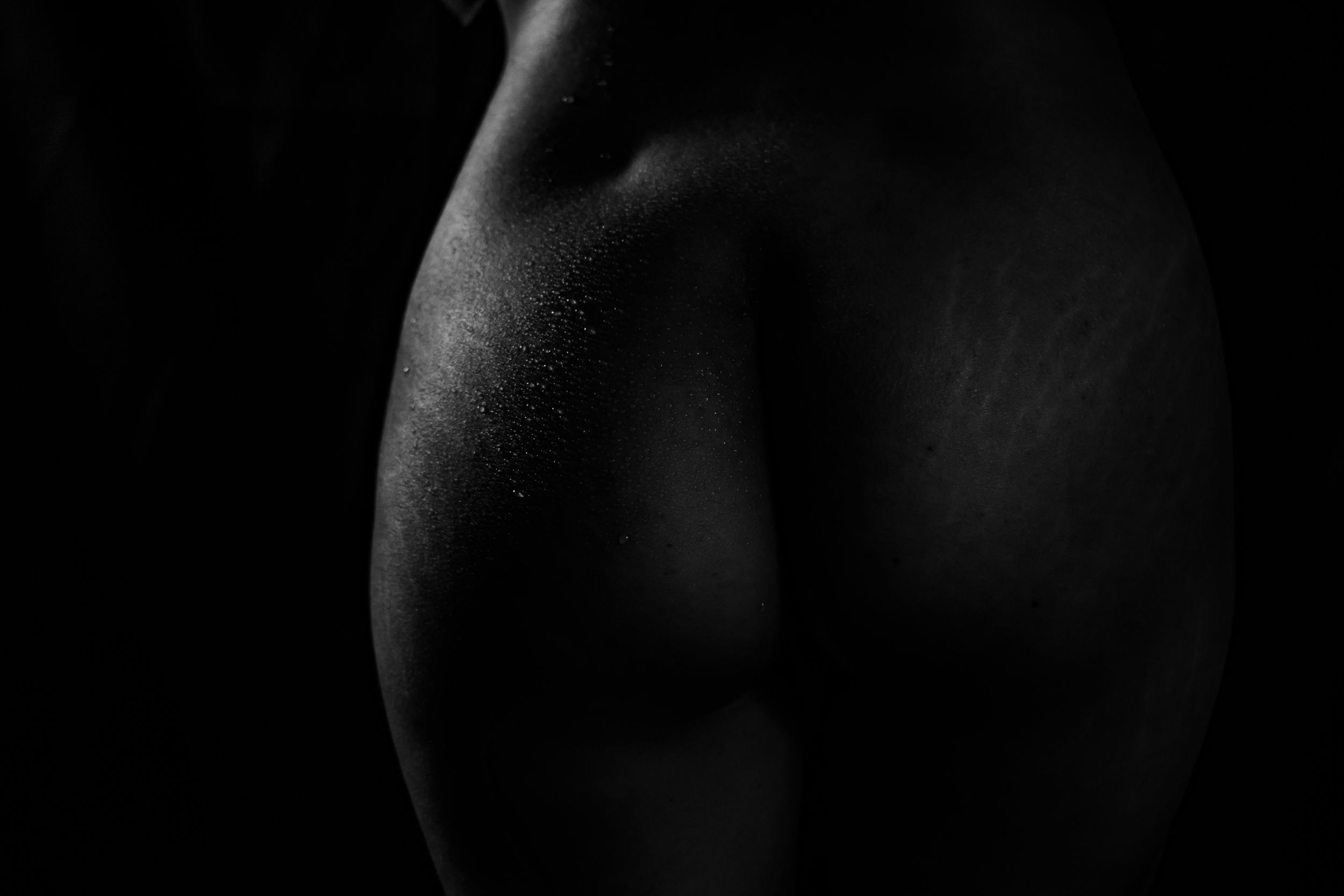 Fotografía artística de curvas femeninas en blanco y negro. Plano detalle de la piel con gotas de agua, iluminación tenue y fondo oscuro.
