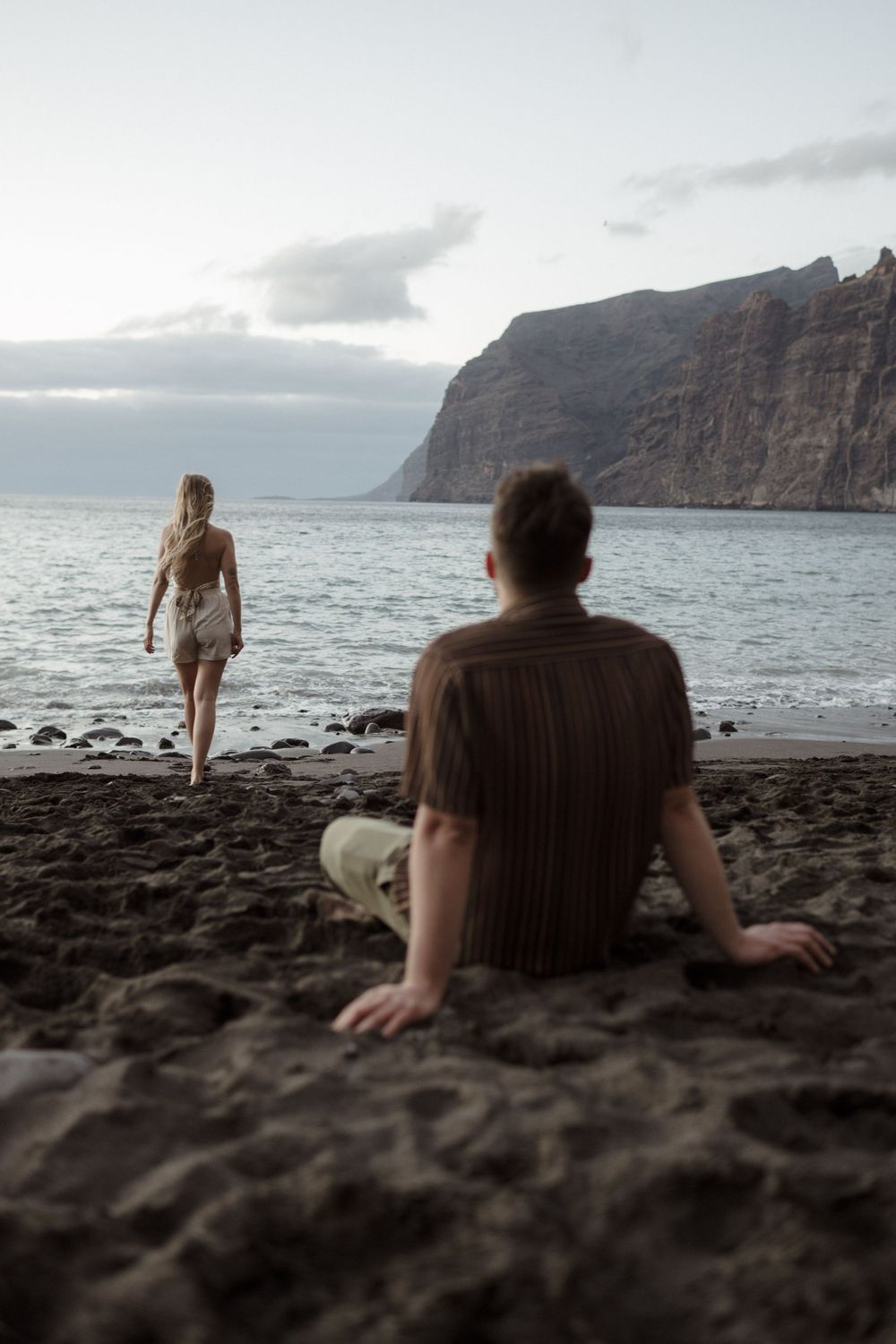 Man sitting on dark sand beach watching woman walk toward ocean with dramatic cliffs in background at dusk.