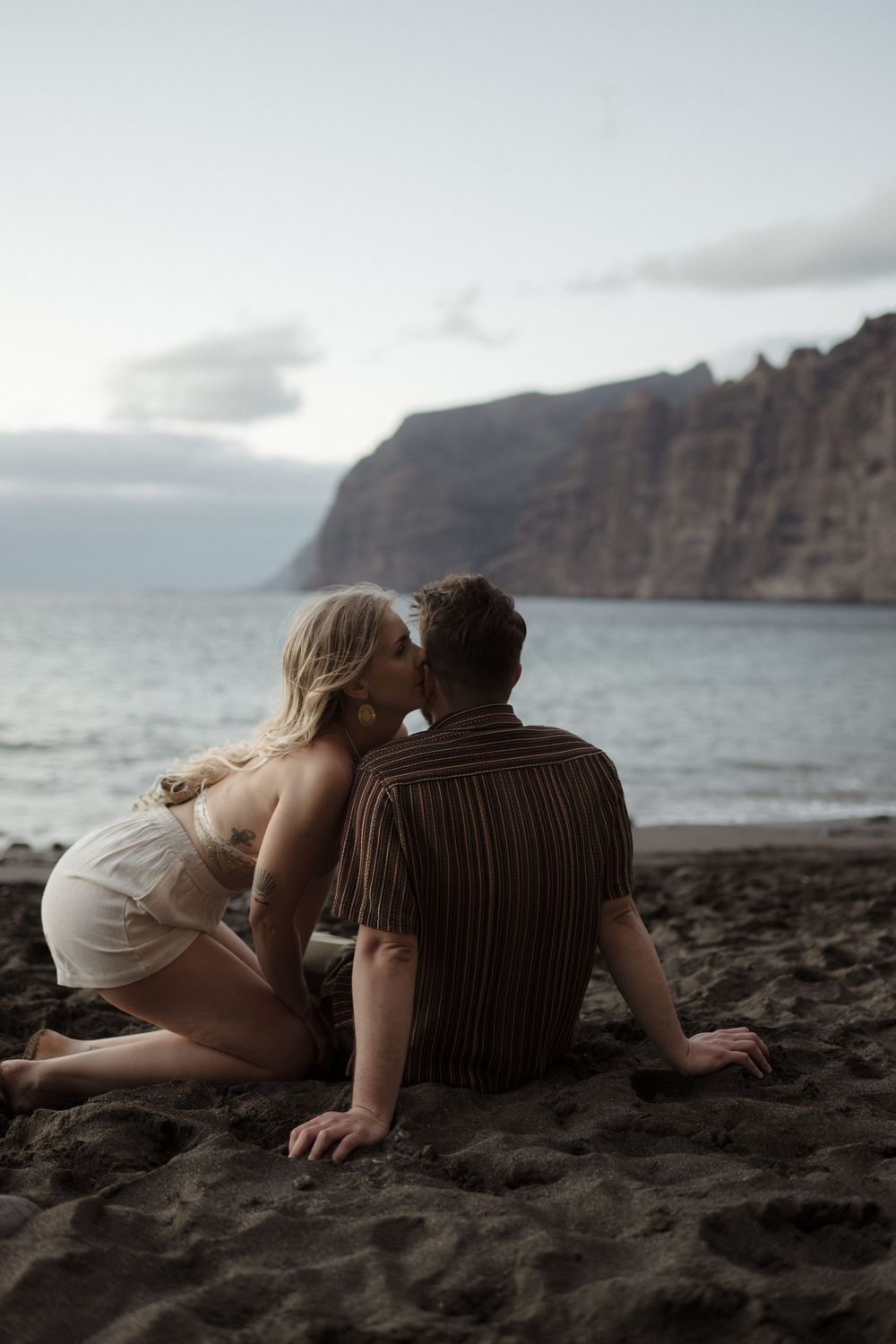 Couple kissing on a dark sandy beach at dusk with dramatic cliffs and ocean in the background.