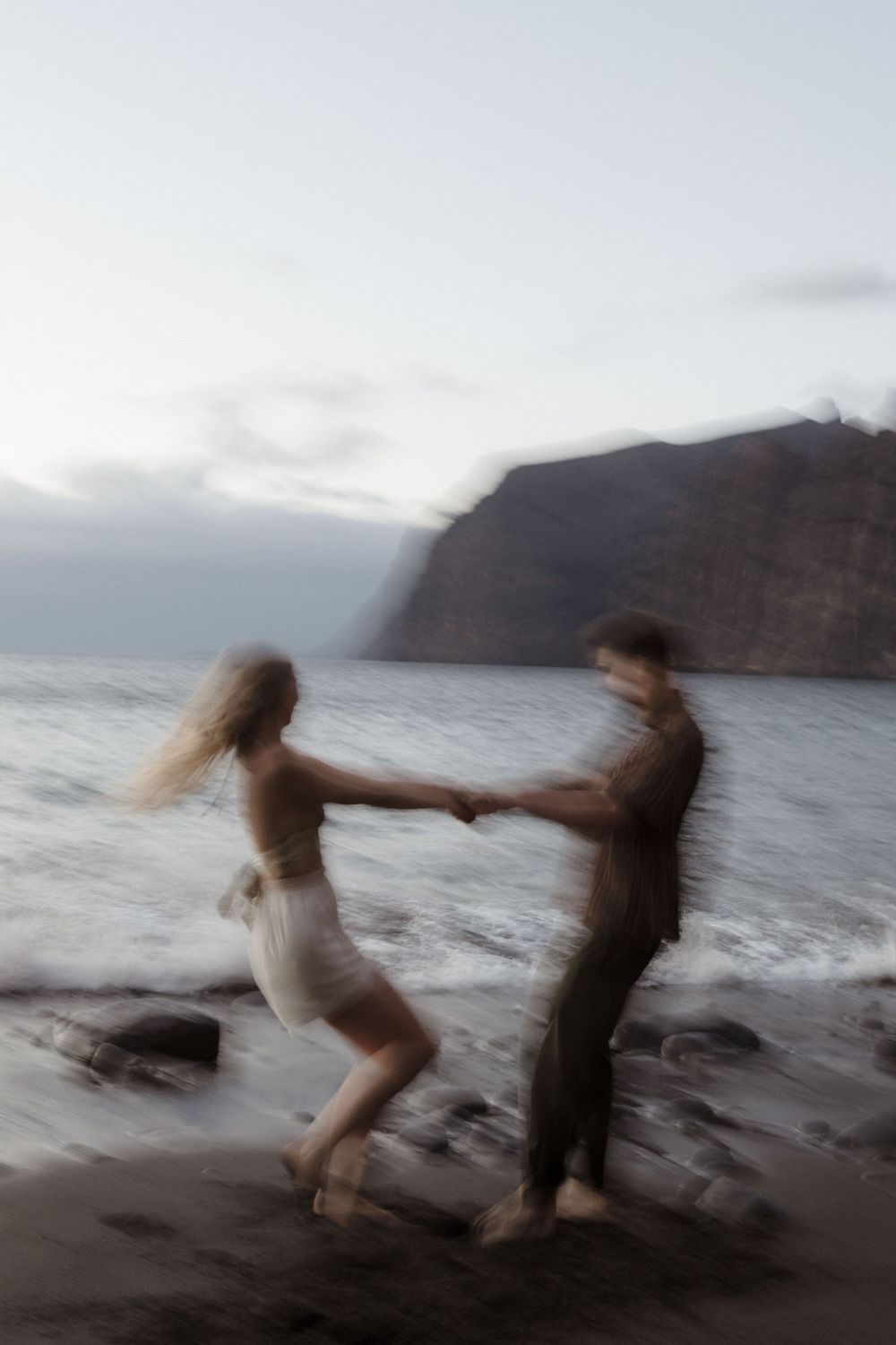 Couple dancing joyfully on a rocky beach at dusk with ocean waves and dramatic cliffs in the background.