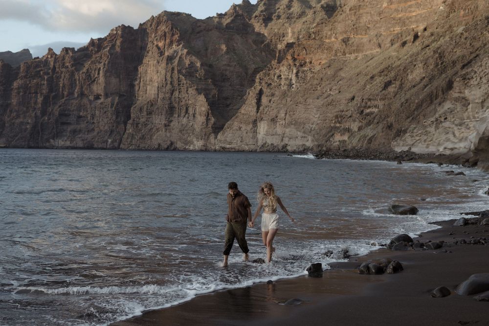 Couple walking hand in hand along a black sand beach with dramatic rocky cliffs in the background at sunset.
