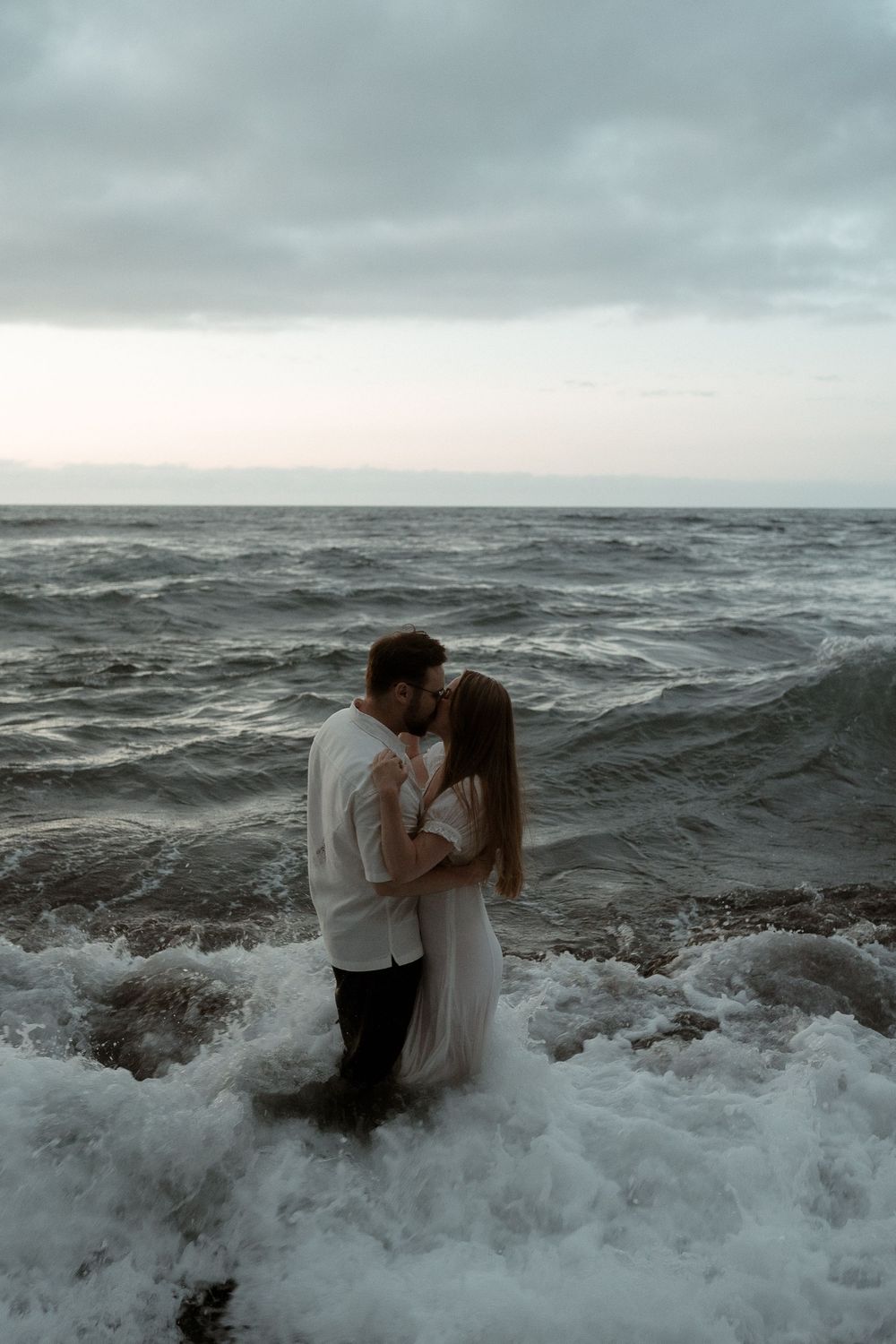 Couple embracing and kissing in ocean waves on a cloudy day, woman in white dress, romantic seaside photo.