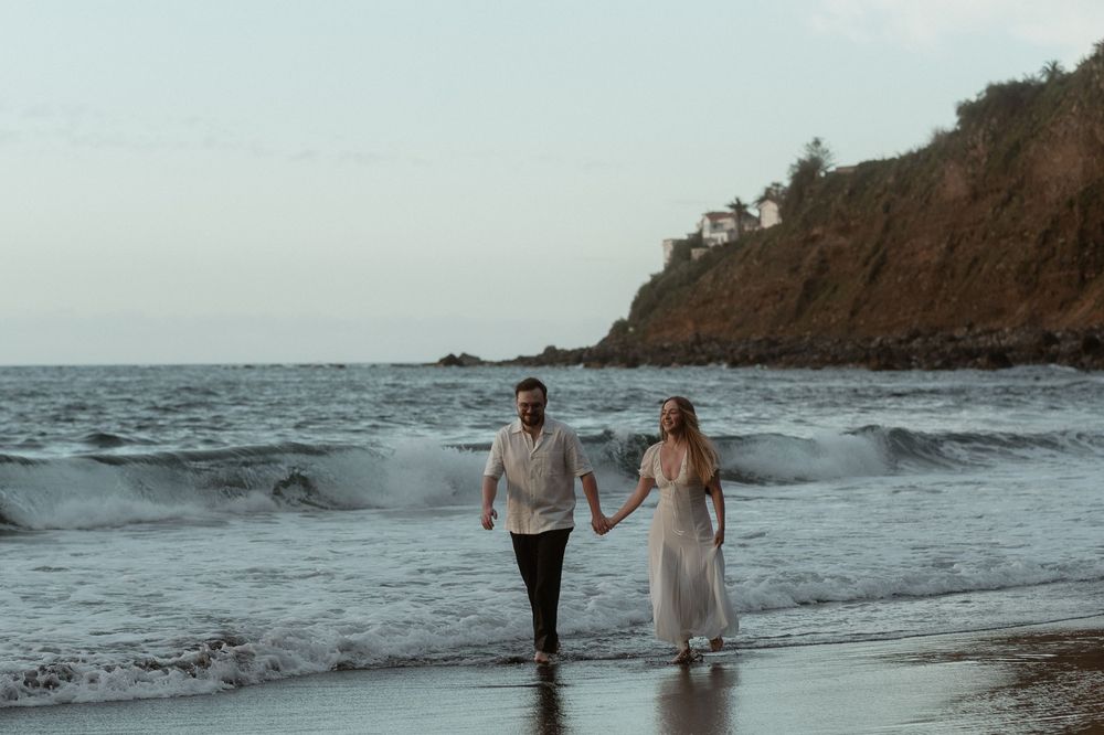 Couple holding hands walking along a scenic beach shoreline with ocean waves and a tree-covered cliff at dusk.