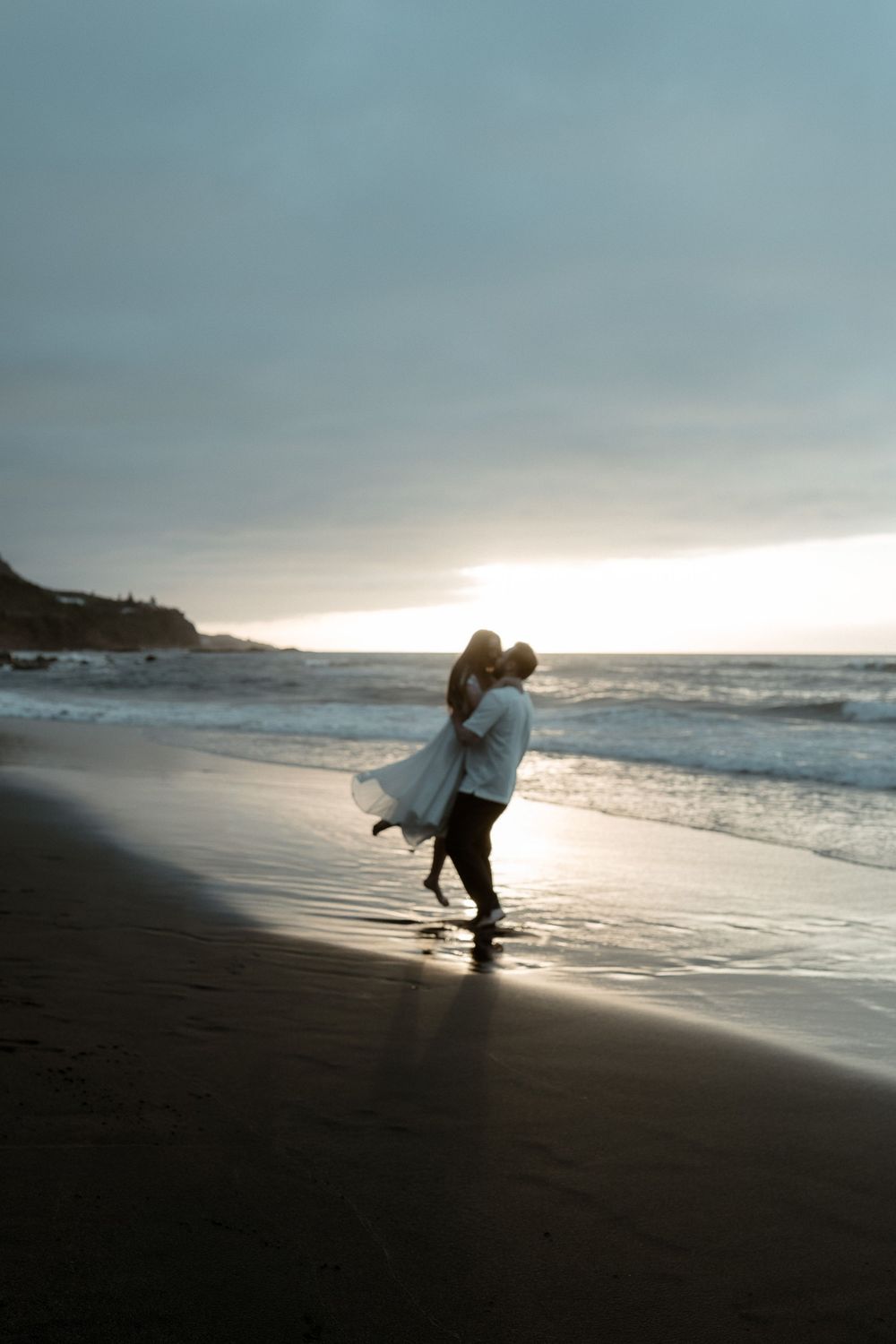 Couple sharing romantic embrace on a sandy beach at sunset, with ocean waves and dramatic cloudy sky backdrop.