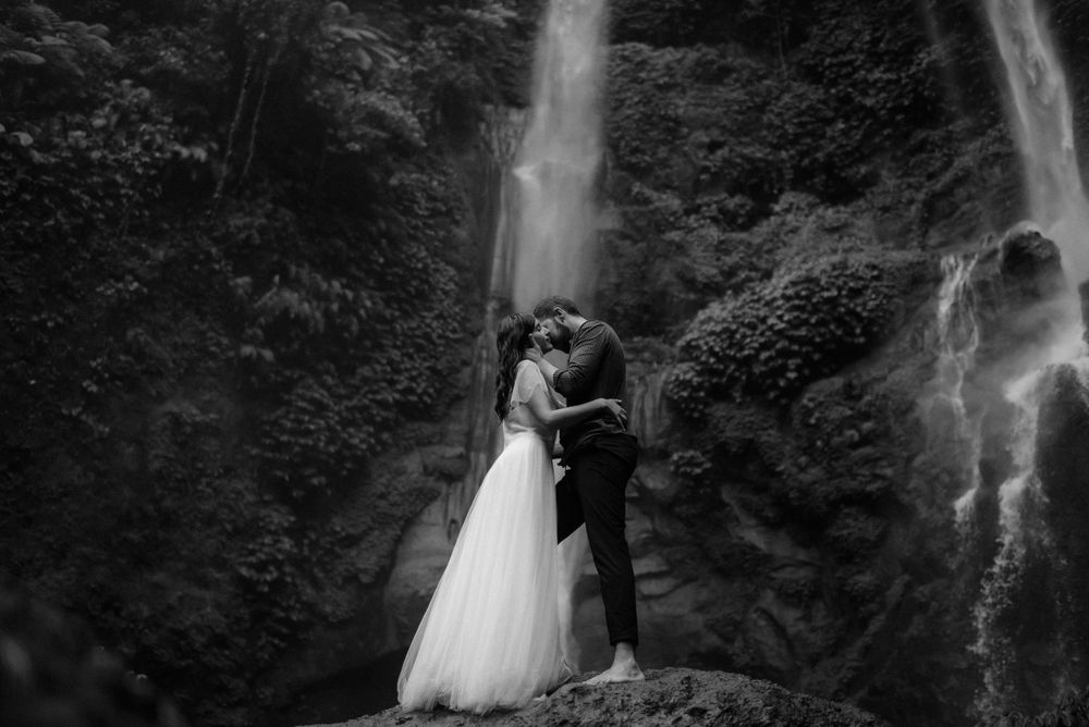 Black and white photo of a couple kissing in wedding attire before a stunning waterfall surrounded by lush greenery.