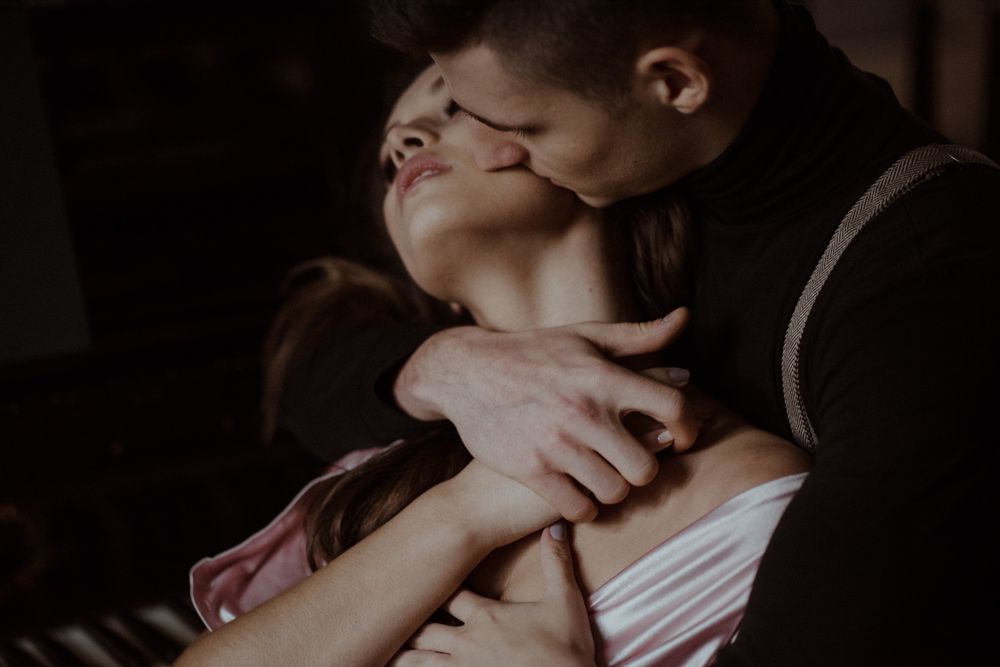 A romantic couple embraces intimately, with a man kissing a woman's forehead in a dark, moody setting.