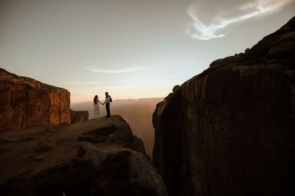 Couple holds hands on rocky cliff edge at sunset with mountain valley views in a dramatic outdoor wedding elopement photo.