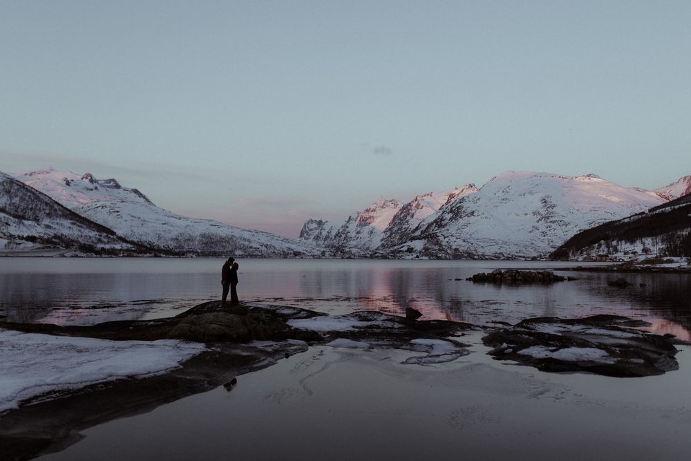 A lone figure stands on rocks beside a serene fjord with snow-capped mountains reflecting in calm waters at dusk.