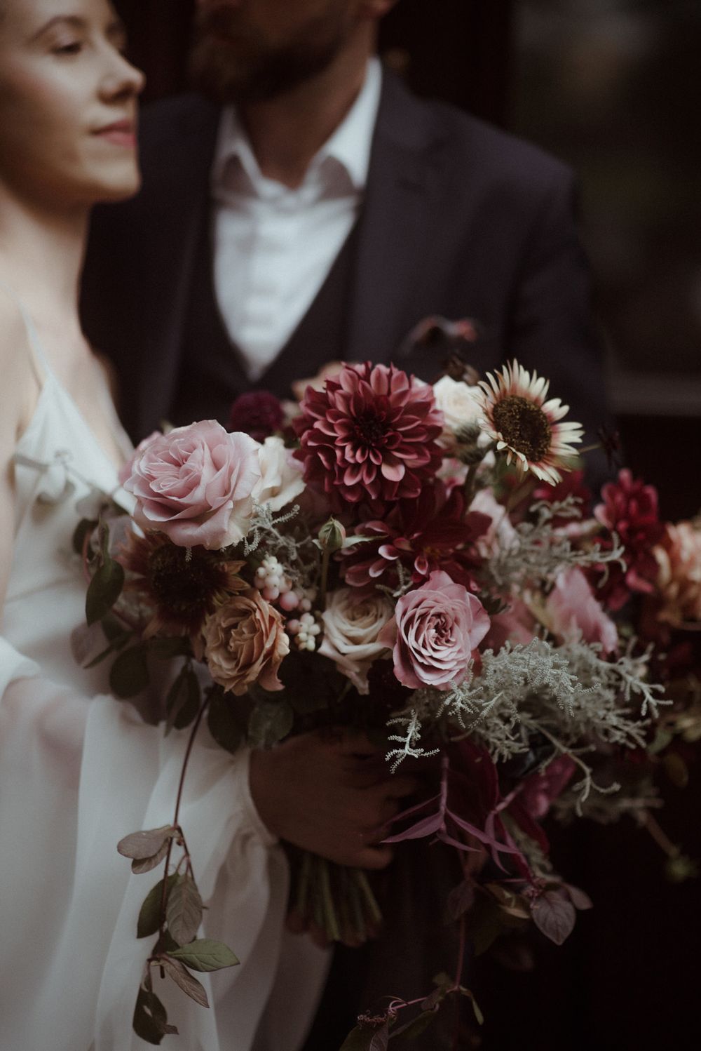 Bride holding a lush wedding bouquet with deep burgundy dahlias, dusty pink roses, and greenery, with groom in dark suit behind her.