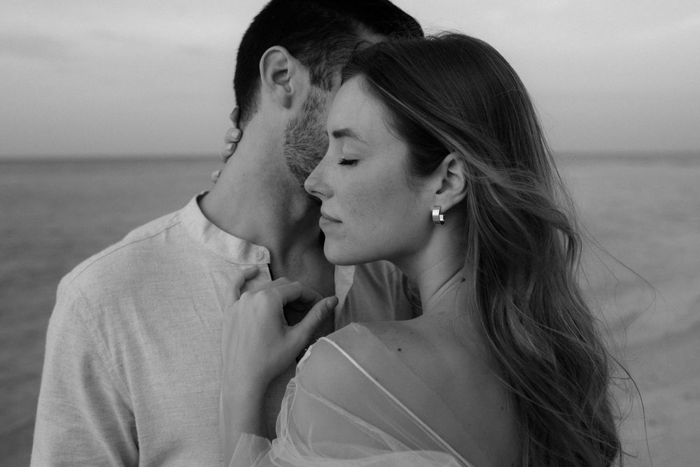Black and white photo of a romantic couple sharing an intimate moment near the ocean at dusk.