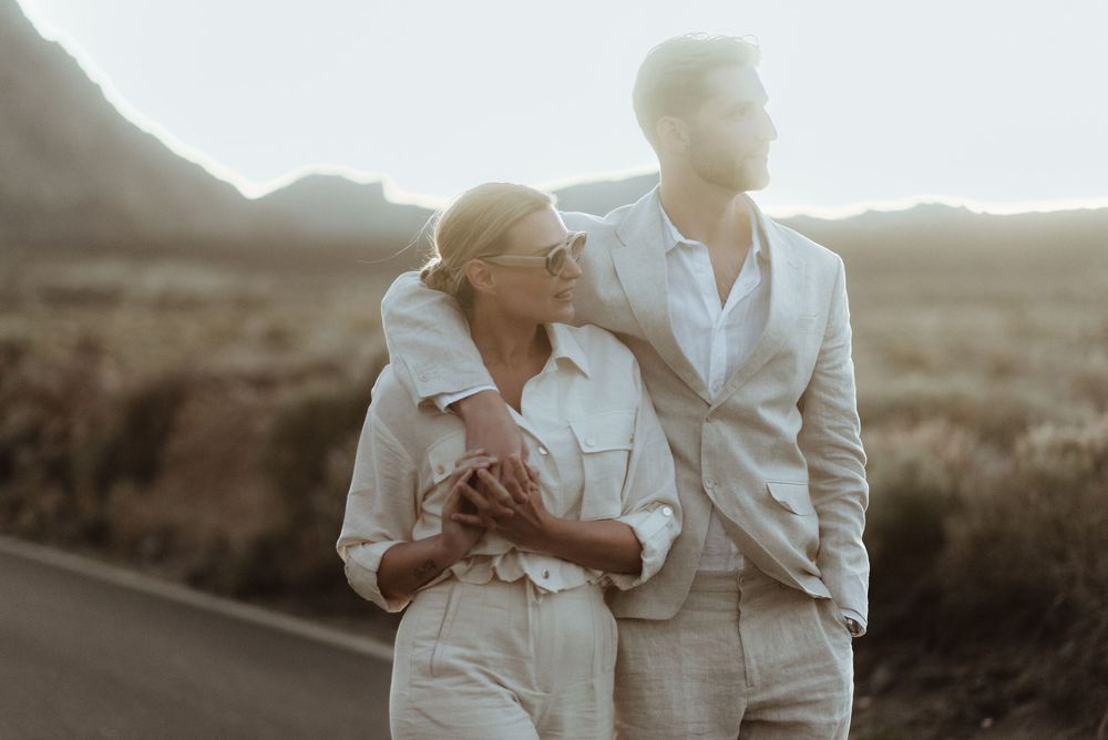 Couple in matching beige outfits embracing on a road with mountains and golden fields in the background.